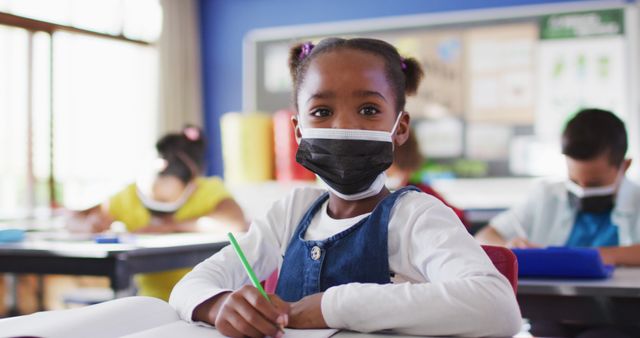 Portrait of african american schoolgirl wearing face mask in classroom looking at camera. children in primary school during coronavirus covid 19 pandemic.