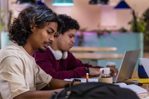 Studying together: indian male friends working on laptops and writing notes in cozy cafe