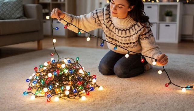 Young woman untangling colorful globe lights in cozy living room wearing knit sweater