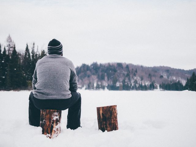 Man is sitting on a wooden log in a snowy landscape, experiencing solitude and serenity. The backdrop features a dense forest with snow-covered ground and overcast sky. This image can be used to depict calmness, introspection, outdoor activities, and the beauty of winter.