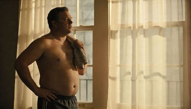 Middle-aged man standing shirtless by sunlit window holding towel, contemplating morning light