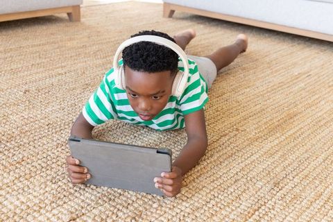 School-age african american boy lying on jute rug using tablet and wearing headphones