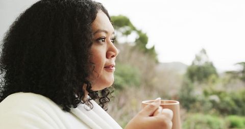 Curly-haired woman in white robe gazing out window holding mug, peaceful morning