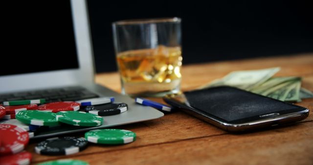 Poker chips and a glass of whiskey are placed next to a smartphone and a laptop on a wooden table, alongside some cash. This photo can be used to depict themes of online gambling, casino activities, and technology integration in gaming. Suitable for articles, advertisements, and blogs related to online casinos, gambling behaviors, and leisure activities.