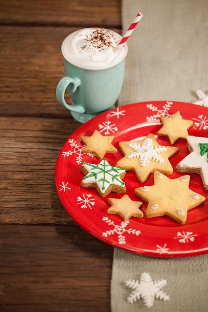 Christmas cookies in various shapes with icing on a red plate, accompanied by a mug of cappuccino on a wooden table. Ideal for holiday-themed designs, winter promotions, and social media posts related to Christmas and festive celebrations.