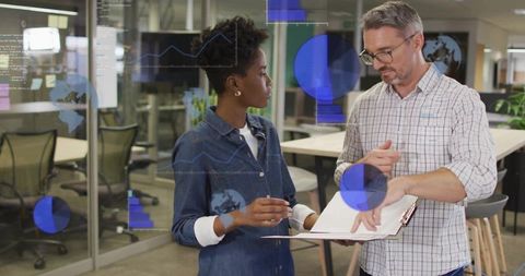 Senior mentor guiding coworker over clipboard with projected charts on glass wall