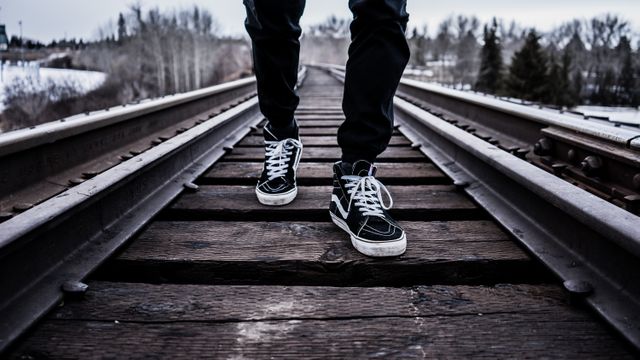 This image portrays a person walking on railroad tracks wearing black sneakers on a cloudy day. The tracks are in a rural countryside setting with trees in the background. This type of image can be used for themes related to adventure, journey, solitude, travel, and exploration. It is ideal for blogs, travel websites, motivational content, and articles that discuss making decisions or taking paths less traveled.