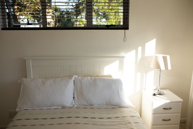 This image showcases a modern bedroom with sunlight streaming through window blinds, illuminating a white bed with pillows and a bed throw. A night table with a table lamp adds to the minimalist decor. Ideal for use in home decor blogs, interior design websites, and advertisements for bedroom furniture or home improvement.
