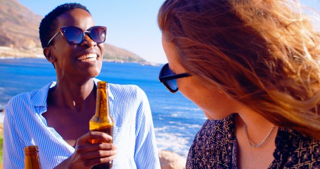 Two friends wearing sunglasses smiling and enjoying beer by the ocean on a sunny day. Ideal for use in travel and leisure advertisements, summer vacation promotions, and social connection themes.