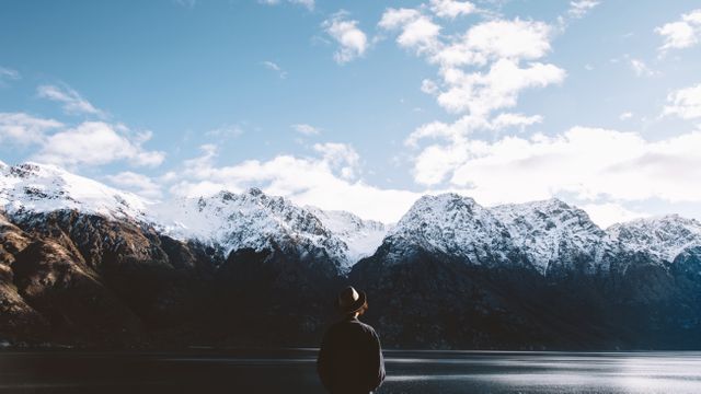 Individual standing near the edge of a calm lake, gazing at a breathtaking view of snow-covered mountains under a partly cloudy sky. Perfect for use in travel blogs, adventure magazines, and promotional material emphasizes nature tourism, adventure travel, and tranquility.