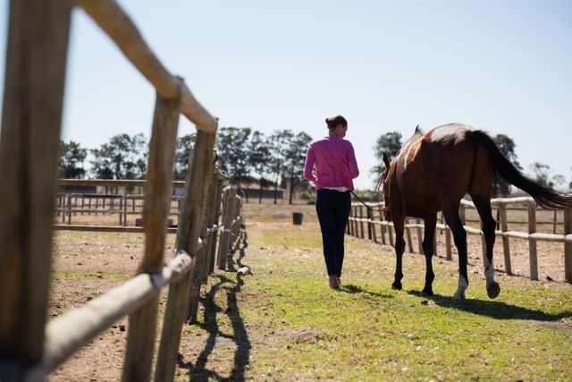 Woman walking with horse at ranch on sunny day, showcasing rural lifestyle and bond between human and animal. Ideal for use in articles about equestrian activities, countryside living, animal care, and outdoor leisure.