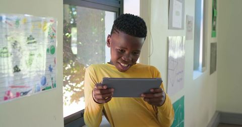 African american teen using tablet in school corridor, leaning on windowsill, smiling