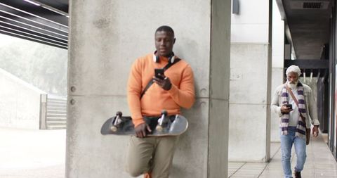 African American man leaning on pillar checking phone with skateboard, senior walking plaza