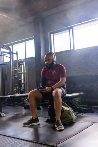 Man sitting on bench listening to music and checking phone at industrial gym post-workout