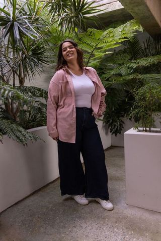 Young woman smiling in indoor atrium with tropical plants, pink overshirt, casual style