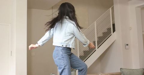 Asian woman jumping on sofa in modern living room swinging hair wearing denim jacket and jeans