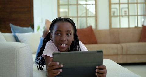 African american preteen girl using tablet lying on ottoman in bright modern living room