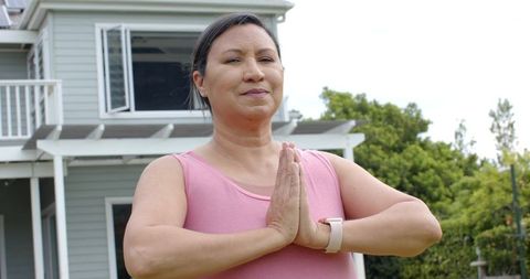 Woman practicing outdoor meditation in front of gray house with smartwatch and pink top