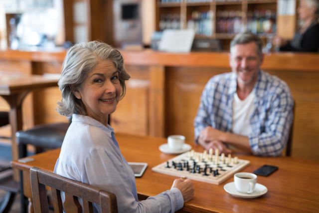 Senior couple enjoying a game of chess in a cozy cafe, both smiling and looking at the camera. Ideal for use in advertisements promoting senior activities, leisure time, and social interactions. Can also be used in articles about healthy aging, mental stimulation, and spending quality time together.