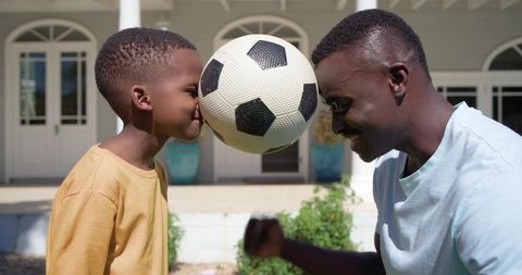 African american father and son pressing soccer ball between foreheads on sunny porch