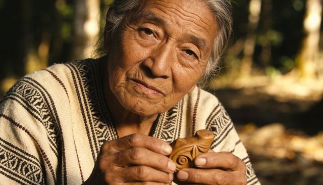 Elderly artisan holding carved wooden figurine in dappled forest light, textured portrait