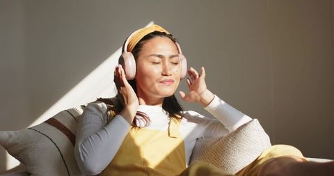 Asian woman relaxing on sofa listening to music with pink headphones in cozy living room