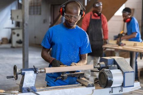 African american woodworker turning lathe while team crafting furniture in carpentry shop