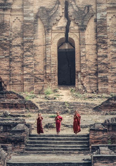Three Buddhist monks in red robes are climbing the weathered stone steps of an ancient temple with a large crack in its facade. Ideal for use in articles or promotions about Buddhism, cultural heritage, and travel in Southeast Asia. This image highlights themes of spirituality, tradition, and architecture.