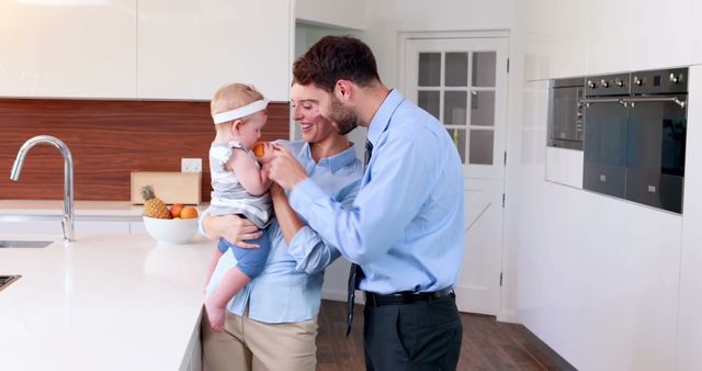 Same-sex couple holding and playing with baby in a bright, modern kitchen. Ideal for use in materials related to family life, LGBT representation, modern parenting, and positive household moments. Useful for advertisements, articles about inclusivity, family brochures, and lifestyle blogs.