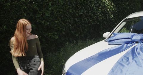 Red-haired woman smiling while receiving new white car with giant blue bow in driveway