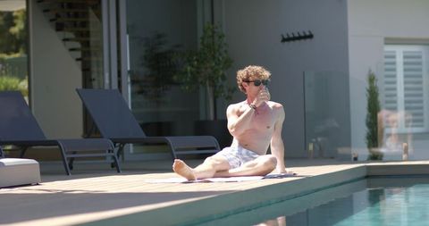 Shirtless man sunbathing on pool deck drinking water wearing sunglasses and swim trunks
