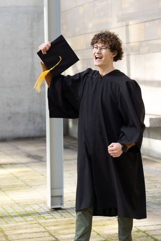 Young graduate raising mortarboard with yellow tassel, clenching fist on sunny campus