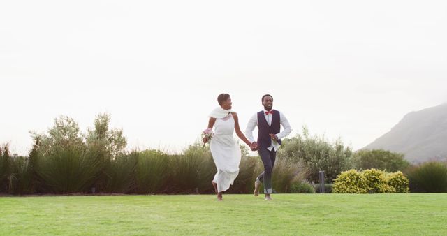 Joyful newlyweds are running hand in hand on a green lawn, surrounded by lush greenery and mountains in the background. Bride is wearing a white dress, and groom is in a suit with a red bow tie. This image conveys happiness, celebration, and the love shared between the couple. Ideal for use in wedding planning websites, romantic blog posts, and wedding invitation designs.