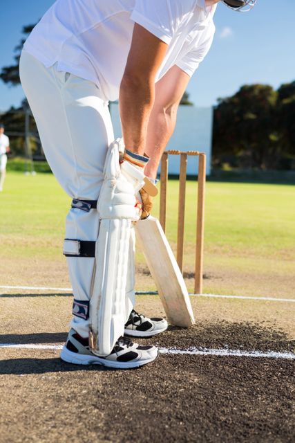 Low section of a cricketer preparing to bat on a sunny day at a sports field. The player is wearing protective gear and holding a cricket bat, ready to face the ball. Ideal for use in sports-related content, fitness promotions, outdoor activity advertisements, and cricket event promotions.