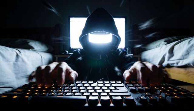 Hooded hacker typing under monitor glow in dark bedroom with orange backlit gaming keyboard