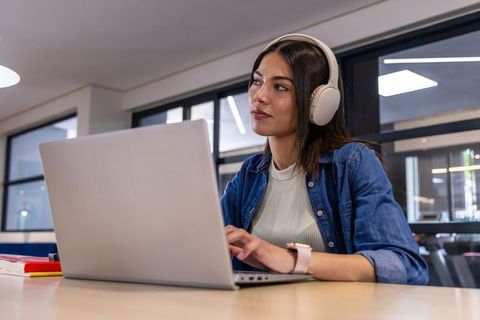 Asian woman wearing headphones working on laptop in modern coworking space, productive