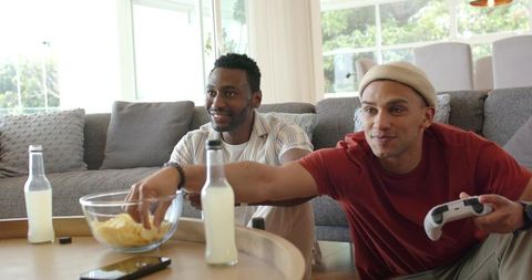 Diverse male friends gaming on couch reaching for chips holding controller and drinks