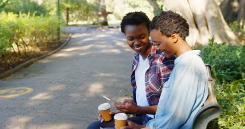 Smiling Twin Sisters Enjoying Coffee, Looking at Phone in Park