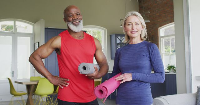 Mature couple standing in loft apartment holding yoga mats, suggesting a healthy and active lifestyle. Ideal for use in promotional materials for fitness programs, wellness retreats, retirement living, and health-related articles.