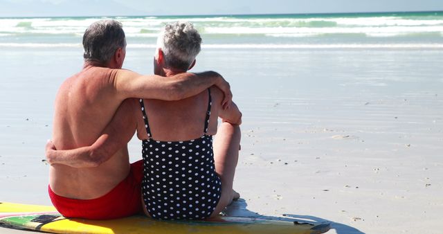Senior couple sitting arm in arm on beach relaxing in vacation atmosphere. They are seated on a surfboard, looking at waves and horizon. Ideal for retirement ads, travel brochures, lifestyle articles, and beach vacation promotions.