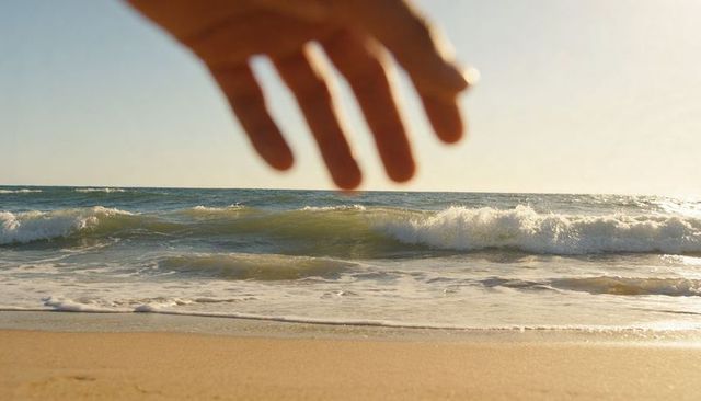 Outstretched hand hovering above sunlit beach shoreline with ring and golden foamy waves