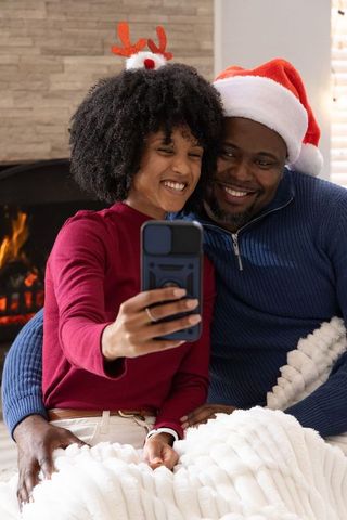 African american couple taking cozy holiday selfie by fireplace wearing santa hats