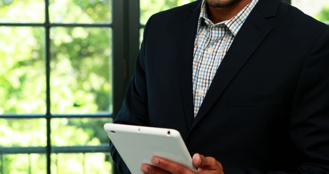 Businessperson in a suit holding and using a digital tablet in an office setting with large windows revealing greenery outside. Ideal for visuals related to business, corporate presentations, workplace productivity, technology in business, and professional settings.
