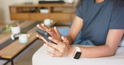 African american woman using smartphone and smartwatch while relaxing on couch at home