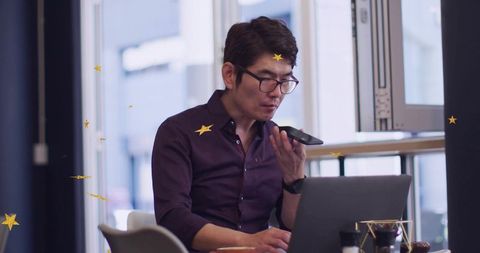Asian professional speaking into smartphone while typing on laptop in urban cafe workspace