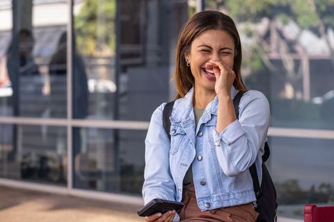 African american woman laughing while using smartphone near urban glass facade