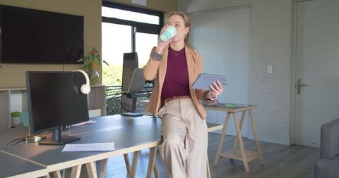 Professional woman sipping travel mug while holding tablet in bright modern home office