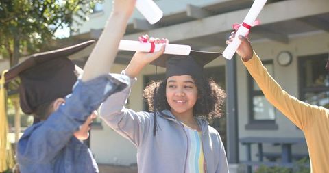 Sunlit diverse students celebrating graduation raising red-ribbon diplomas and mortarboards outdoors