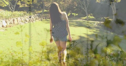 Young woman walking barefoot through sunlit meadow with wildflowers and stone wall