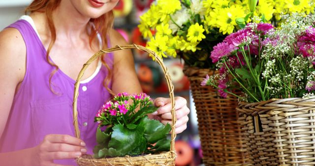 This stock photo shows a woman arranging a basket filled with flowers in a floral shop. The woman, dressed in a purple top, focuses on the pink blossoms as baskets filled with vibrant yellow and pink flowers surround her. This image can be used to depict activities related to gardening, floristry, spring decorations, and home decor ideas. Perfect for blogs, websites, and marketing materials related to flower shops and gardening enthusiasts.